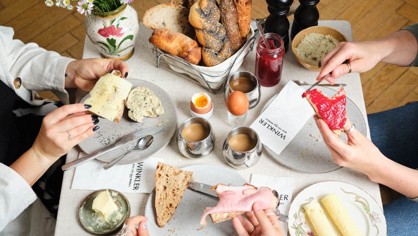Bäckerei Winkler Frühstückstisch mit Gebäck und Händen die Besteck halten und Servietten mit Bäckerei Branding für Bäckerei Winkler